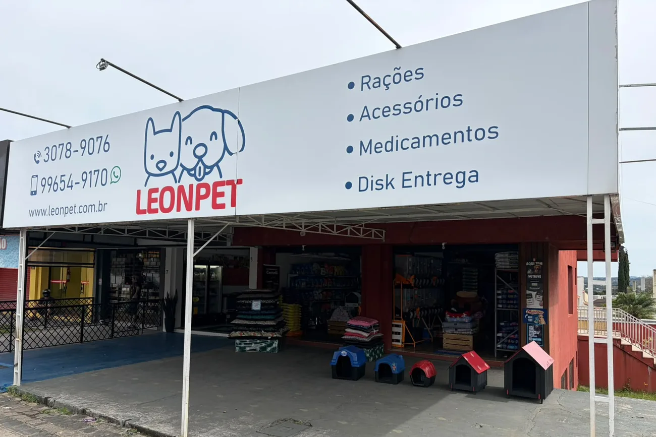 A loja LeonPet vista da rua: a placa branca com o logo, os telefones 3078-9076 e 99654-9170, o endereço www.leonpet.com.br e a lista Rações, Acessórios, Medicamentos e Disk Entrega; embaixo, a loja aberta, prateleiras cheias e casinhas de cachorro na calçada