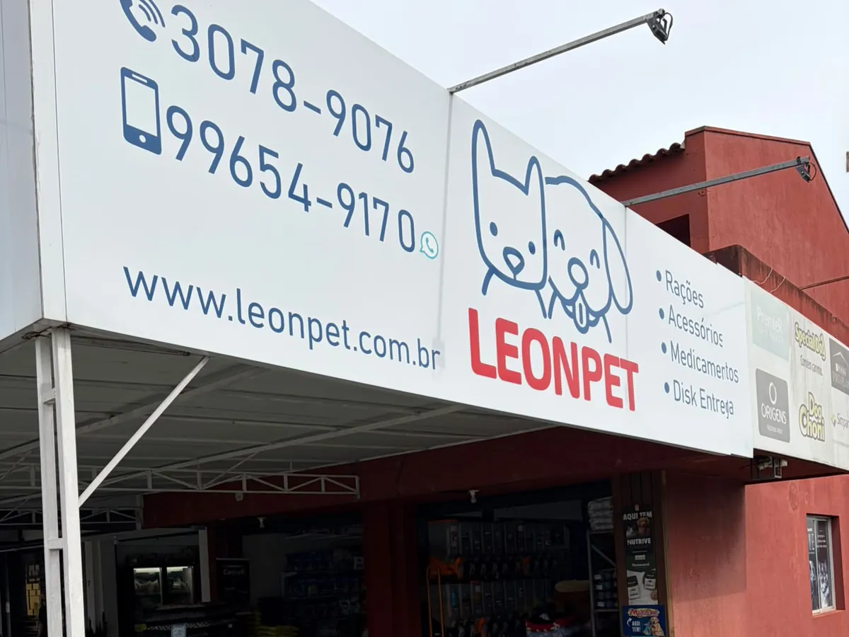 A frente da loja LeonPet vista de baixo: a placa branca com os telefones 3078-9076 e 99654-9170, o endereço www.leonpet.com.br, o logo e a lista Rações, Acessórios, Medicamentos e Disk Entrega; embaixo, a loja aberta com as casinhas de cachorro na entrada